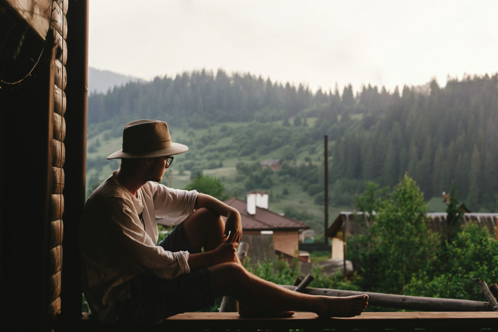 Hipster,Man,Sitting,On,Porch,Of,Wooden,House,Looking,At